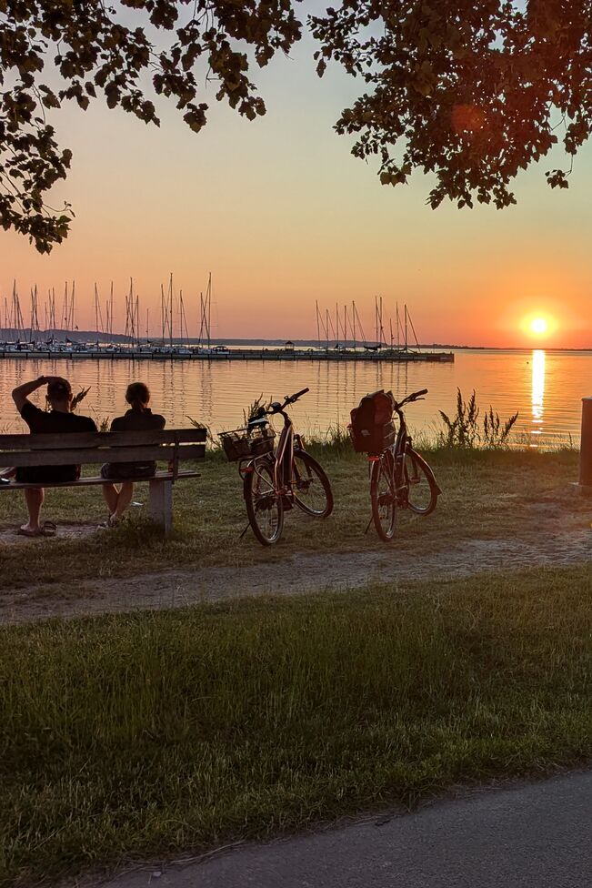 Paar sitzt bei Sonnenuntergang auf einer Bank am Hafen, daneben zwei Fahrräder